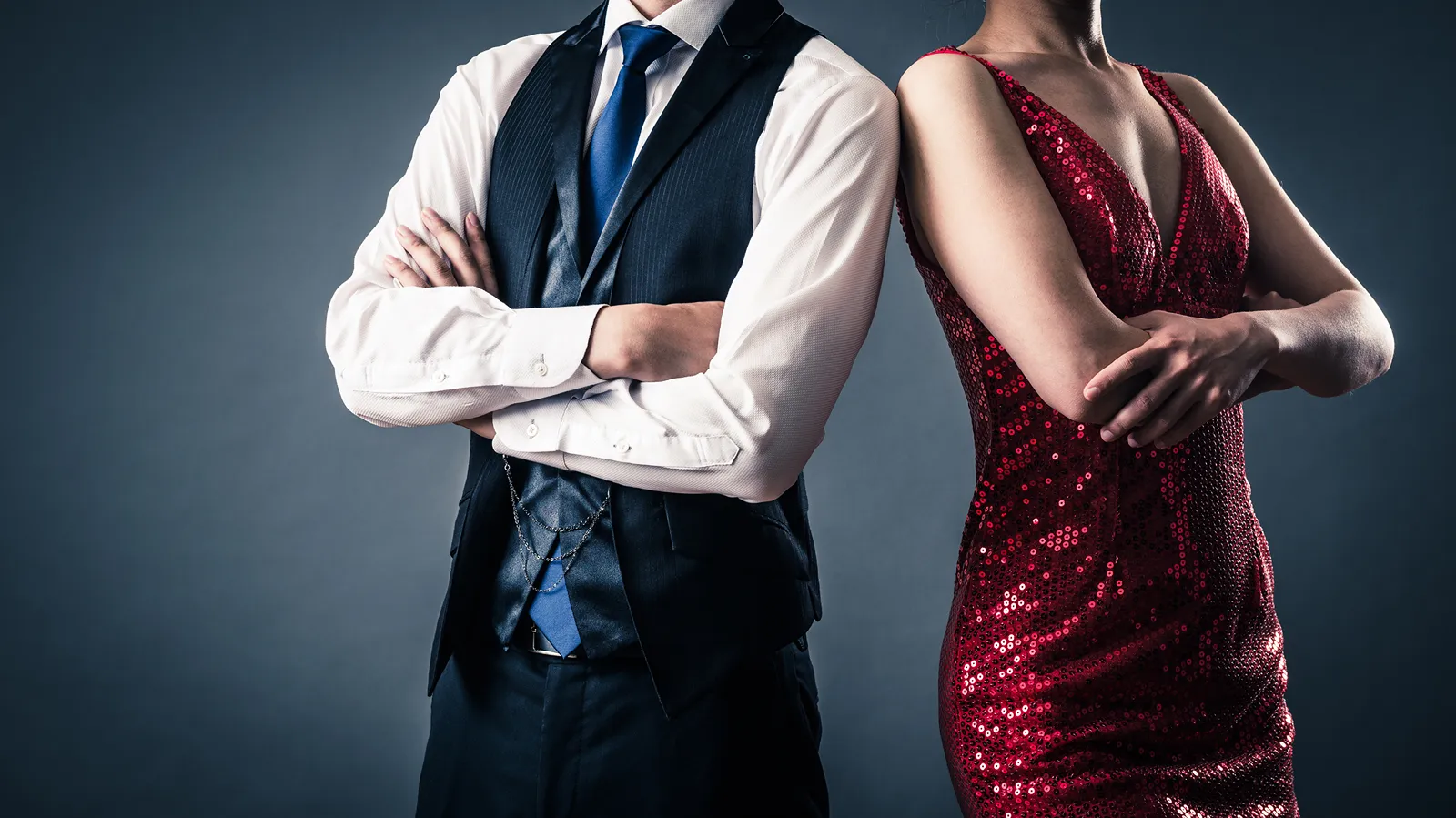 A man in a navy waistcoat and a woman in a red sequinned dress stand back to back with folded arms, both cropped below the shoulders, against a grey background.