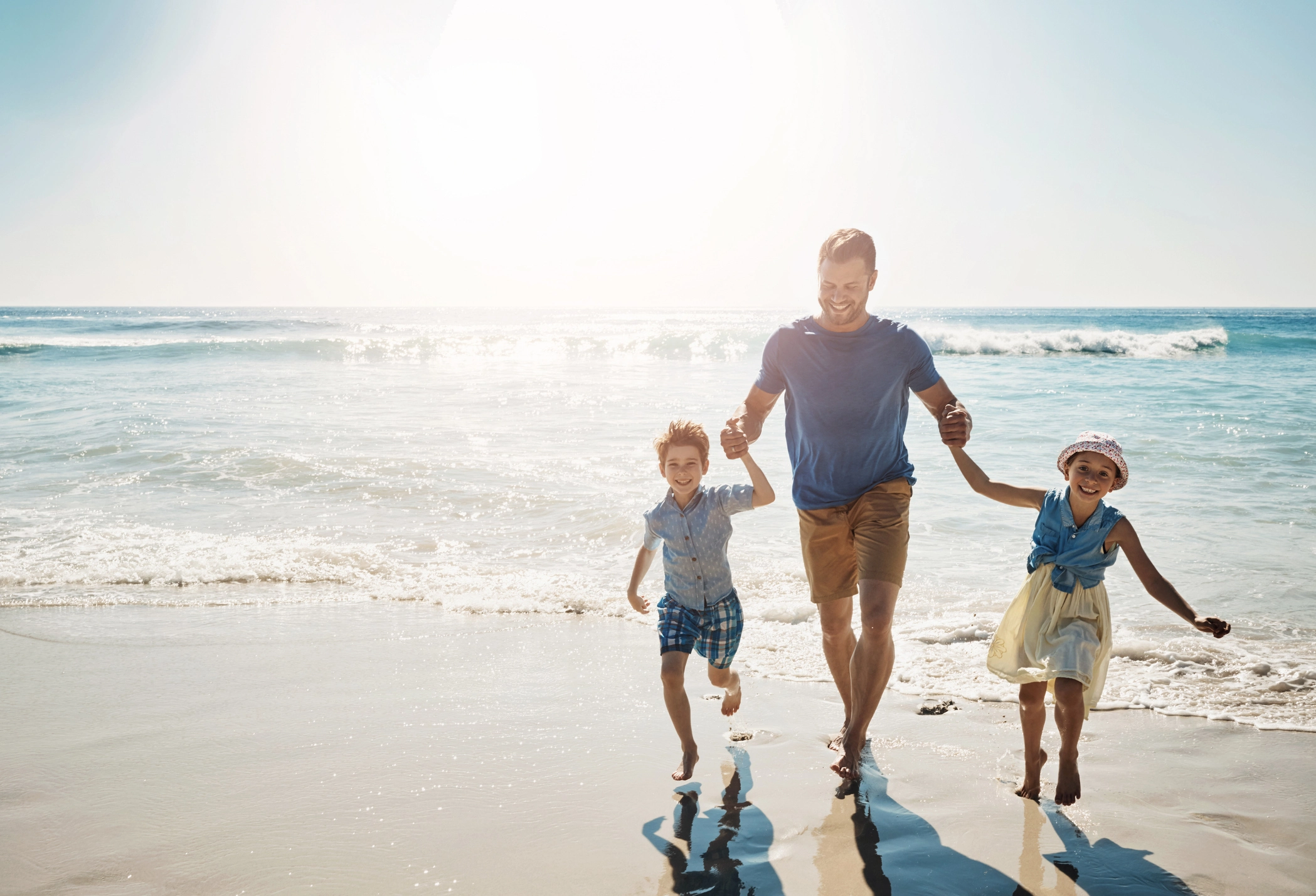 Father holding hands with his son and daughter as they run along a beach on an overseas holiday.