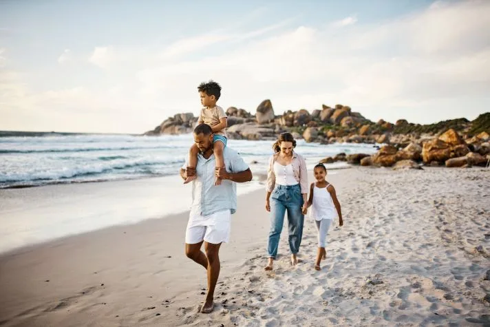 A family of four walks along a sandy beach at sunset, with a father carrying a young child on his shoulders while a mother and daughter follow behind near the shoreline before starting divorce proceedings.