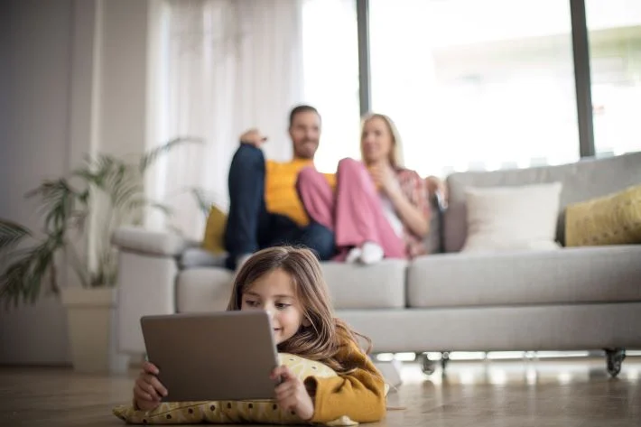 Little girl plays on digital tablet as parents talk on sofa.