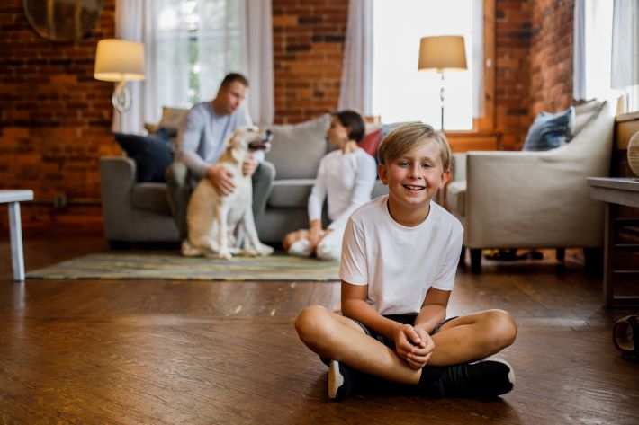 Boy sitting on the floor, joyfully smiling at camera, his parents and dog bonding in the background.