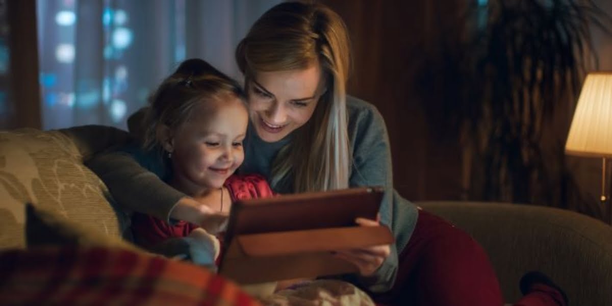 Mother and young daughter are sitting on the sofa at home in the evening, playing on a tablet computer.