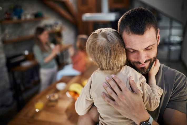 A content father embraces his small son at home while his wife and daughter play in the background.