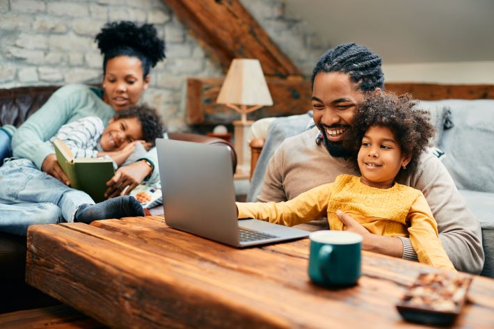 A father and daughter play on a laptop while the mother and son read.